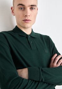Polo de lungă manecă, de culoare verde închis, confecționat din bumbac. Are un guler clasic, plachetă cu trei nasturi și un mic patch cu un logo negru pe piept.