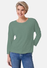 Grünes, strukturiertes Langarmoberteil mit rundem Ausschnitt und abgesenkten Schultern, kombiniert mit blauen Jeans. Glattes, leichtes Material.