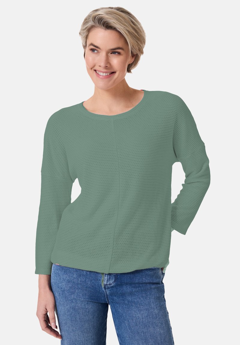 Grünes, strukturiertes Langarmoberteil mit rundem Ausschnitt und abgesenkten Schultern, kombiniert mit blauen Jeans. Glattes, leichtes Material.