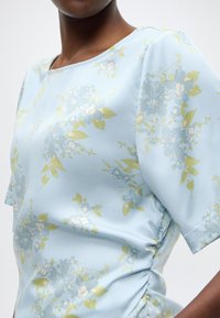 Ανοιχτό μπλε τοπ με κοντά μανίκια και λουλουδάτο σχέδιο σε πράσινο και μπλε. Λείο ύφασμα με συγκεντρωμένες λεπτομέρειες στο πλάι.