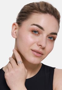 Anello d'argento con bande intrecciate e piccole pietre trasparenti. Il modello indossa una maglietta dai toni neutri e tocca il proprio collo con una mano.
