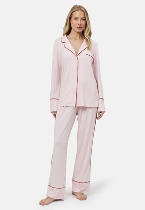 Kobieta stoi boso, ubrana w bladoróżowy, długi rękaw, zapinany na guziki top do piżamy oraz pasujące spodnie z czerwonym obszyciem wzdłuż krawędzi.