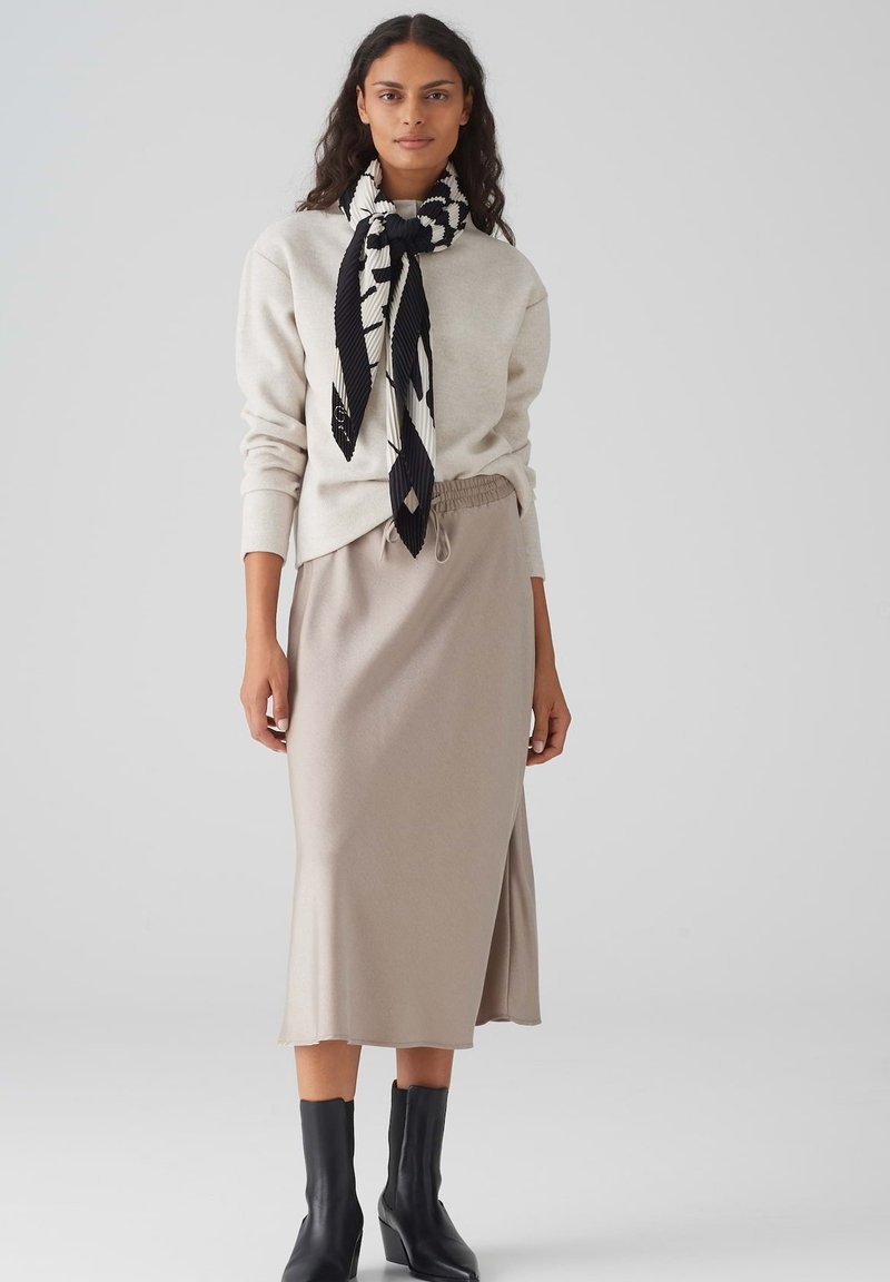 Light gray sweater, beige skirt with an elastic waist, and black ankle boots. Accented with a patterned black and white scarf tied at the neck.