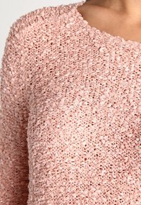 Gros plan sur un pull tricoté rose pâle texturé porté par une personne, mettant en valeur le motif bouclé et tissé du tissu.