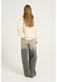 Kobieta z długimi blond włosami, ubrana w kremowy sweter i wyblakłe szare spodnie szerokie, stojąca z jedną ręką w kieszeni, odwrócona plecami.