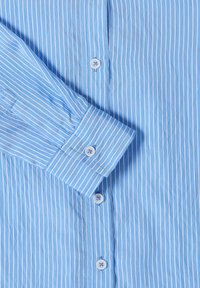 Chemise bleue à rayures verticales blanches, boutonnée à l'avant, avec une manche pliée sur la poitrine montrant le poignet et le bouton.