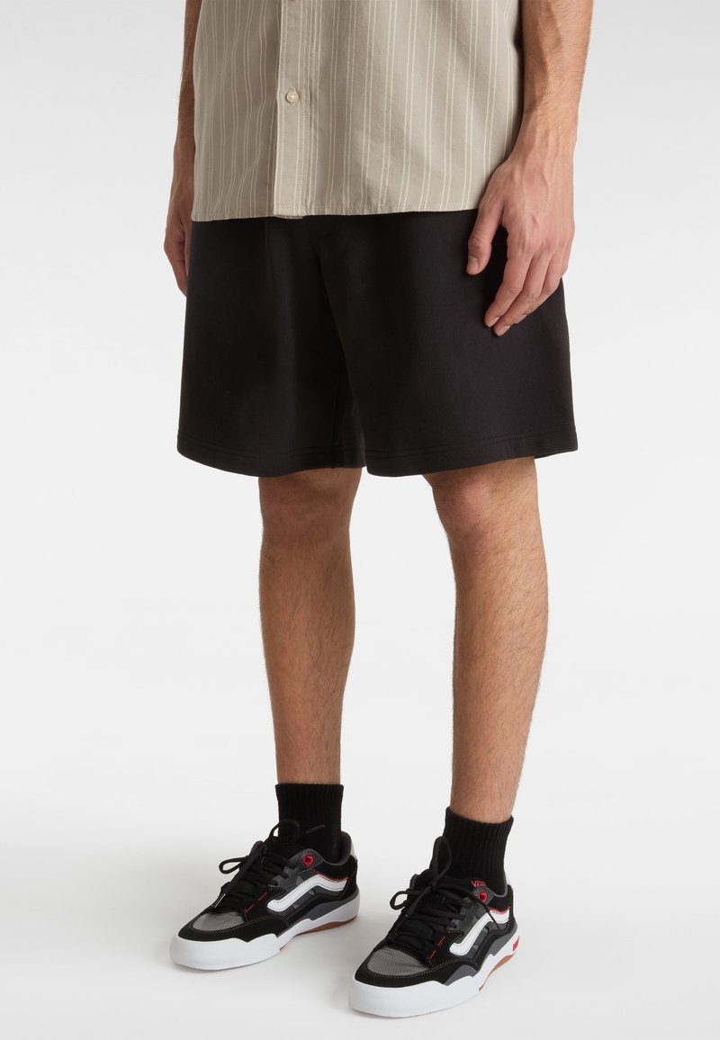 Homme portant une chemise beige rayée, un short noir, des chaussettes noires et des baskets noir et blanc, debout sur un fond blanc.