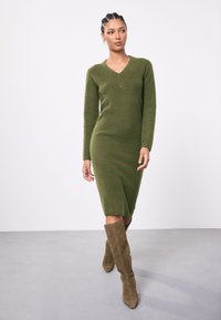 Vestido de punto verde oliva con escote en V, mangas largas, textura acanalada y longitud hasta la rodilla. Combinado con botas marrones de ante hasta la rodilla.