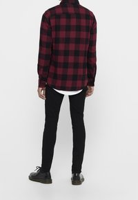 Chemise flanelle à carreaux rouges et noirs avec des manches longues, ourlet incurvé, portée par dessus un T-shirt blanc. Associée à un pantalon noir slim et des chaussures noires.