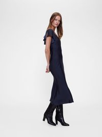 Rochie din satin bleumarin cu mâneci volan, fit slim, până la genunchi. Pur și simplu asortată cu cizmulițe cu toc negre, pe un fundal alb simplu.