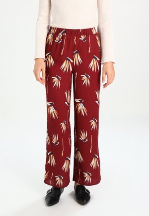 Kobieta w bordowych spódnicowych spodniach z wysokim stanem i szerokimi nogawkami, ozdobionych beżowymi i granatowymi abstrakcyjnymi kwiatowymi wzorami, w czarnych spiczastych wsuwanych butach oraz w beżowej, prążkowanej bluzce z długim rękawem.