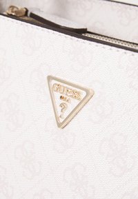Borsa a tracolla bianca con texture, caratterizzata da una placca triangolare dorata con la scritta "GUESS" e un punto interrogativo, chiusura con zip e un delicato motivo in rilievo.