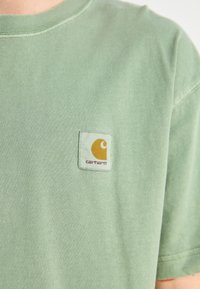 Light green cotton t-shirt with a crew neck and short sleeves. Features a square label with a yellow and orange logo on the chest pocket.