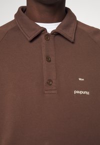 Polo marrone con finitura testurizzata, tre bottoni scuri, logo bianco e testo che recita "Won" e "Hundred" sul lato sinistro.