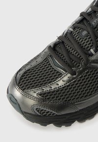 Svart idrottssko med nätöverdel, glänsande syntetiska detaljer, rund tå och släta svarta snören. Mönstrad sula för grepp.