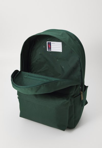 Mochila de tela verde con una apertura grande, bolsillo frontal, cierres de cremallera y una etiqueta interna para la identificación de propiedades.