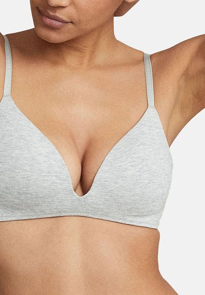 Femme portant un soutien-gorge sans couture gris clair avec des bretelles fines réglables et un décolleté en V profond sur un fond neutre.