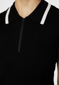 Black ribbed top with a zippered neck, white striped accents on the collar, and a fitted design. Features a V-shaped seam detail.