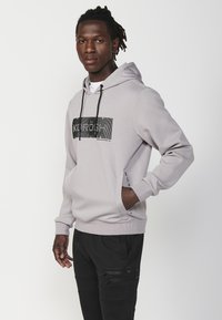 Felpa grigia chiara con una stampa grafica nera sul davanti, cappuccio con cordino regolabile e tasche laterali. Abbinata a pantaloni jogger neri.