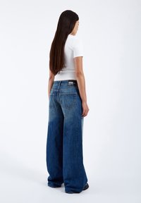 Wide-leg jeans in faded blue denim, featuring a high waist, two back pockets, and a black brand label at the waistband, paired with a white top.
