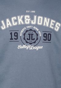 Maglietta in cotone blu con stampa grafica bianca e navy. Il design include i testi "JACK&JONES," "EST 1990," e "College League" con un logo circolare.