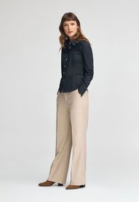 Camicia nera con bottoni e un piccolo logo rosso, abbinata a pantaloni beige larghi e scarpe marroni. Fit slim e texture del tessuto liscia.