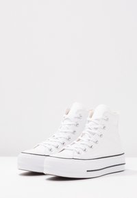 Pair of white high-top canvas sneakers with white laces and thick white rubber soles with black trim, on white surface.