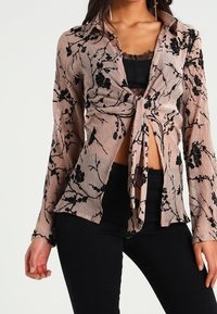 Blusa a maniche lunghe con nodo davanti in tessuto trasparente color pesca con motivi floreali neri. Caratterizzata da un colletto e una superficie testurizzata. Abbinata a pantaloni neri.