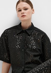 Zwarte kanten shirt met korte mouwen, voorzien van bloemen uitsnijdingen, een kraag en knopen, gemaakt van een gestructureerde stof met een semi-transparant ontwerp.