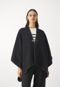 Svart ullponcho med buet kant, med knappelukking foran og kontraststingdetaljer. Brukt over enstripete topp.
