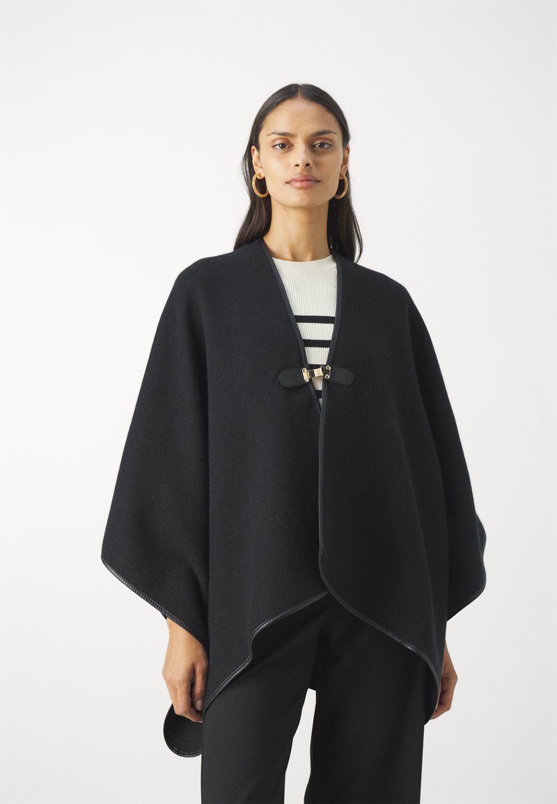 Svart ullponcho med buet kant, med knappelukking foran og kontraststingdetaljer. Brukt over enstripete topp.