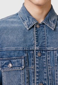 Jeans jakna s klasičnom kragnom, dvije prednje džepove, metalnim kopčama i svijetloplavim izblijedjelim završetkom s suptilnom teksturom.