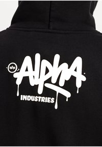 Μαύρο φούτερ με μεγάλο λευκό λογότυπο «Alpha Industries» σε στυλ γκράφιτι στην πλάτη, με σταγόνες και μικρό στρογγυλό εικονίδιο.