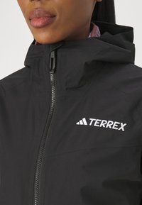 Black Terrex jacket with a high collar, front zipper, and embroidered logo. Smooth texture with a matte finish and minimalistic design.