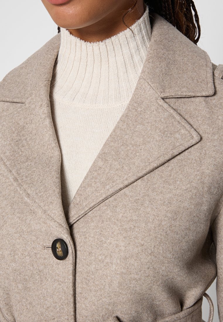 Cappotto beige in misto lana con rever a punta, chiusura a un bottone e maglione bianco a coste testurizzato sotto. Tessuto morbido e caldo.