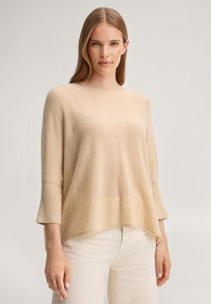 Vrouw met steil, lang blond haar draagt een losse beige trui met geribde boorden en off-white broek, staand tegen een effen achtergrond.