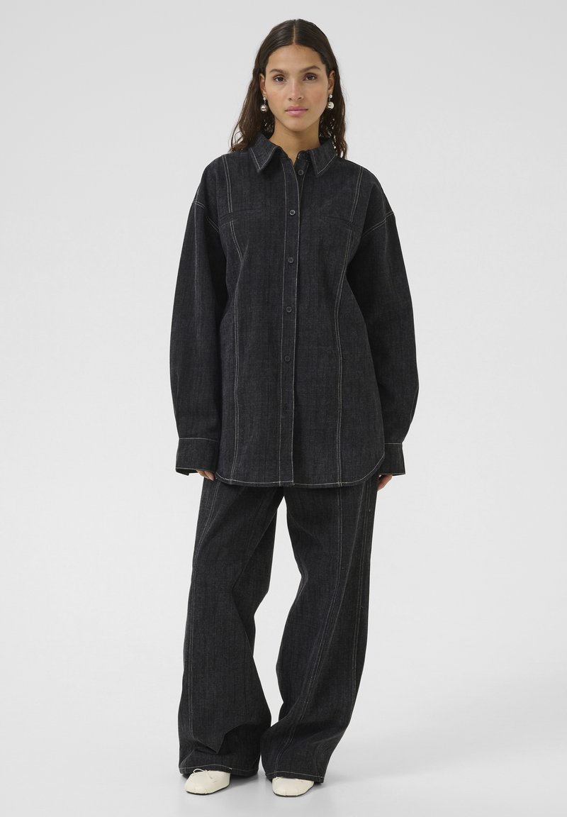 Tumšs džinsa divdaļīgs komplekts, kas ietver pogājamu krekla ar klasisku apkakli un platiem biksēm. Kontrasta šuvumi izceļ dizainu.