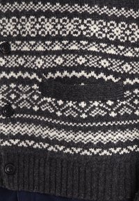 Cárdigan de punto gris oscuro con patrones geométricos blancos, que presenta un dobladillo acanalado y un pequeño bolsillo en el pecho, cerrado con botones negros.