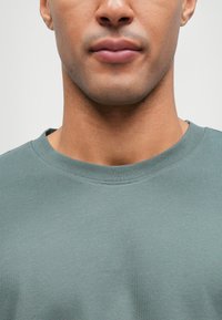 Πράσινο βαμβακερό φούτερ με στρογγυλή λαιμόκοψη και υφή υφάσματος, σε κλασική γραμμή. Η λαιμόκοψη είναι ραμμένη, αναδεικνύοντας τα στοιχεία της χαλαρής σχεδίασης.