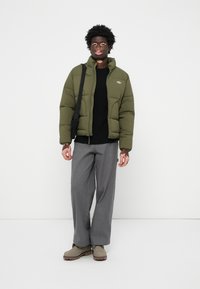 Olivengrøn pufferjakke med høj krave, sammen med en sort strikket sweater, grå vide bukser og beige ankelstøvler.