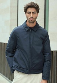 Jachetă bleumarin, cu fermoar, cu guler, buzunare laterale și un finisaj neted, texturat. Design modern cu o siluetă ajustată.