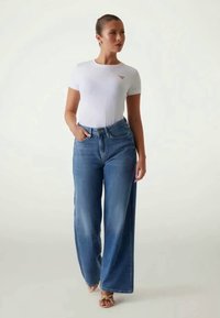 T-shirt bianco aderente con un piccolo logo, abbinato a jeans in denim blu a vita alta e larghi. La modella indossa sandali beige.