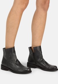 Bottes cheville en cuir noir à texture lisse, fermeture éclair latérale et talon légèrement surélevé. Caractérisées par un design minimaliste et une tige découpée.