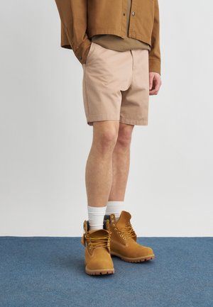 Mężczyzna ubrany w beżowe szorty, brązową kurtkę, białe skarpetki i jasnobrązowe buty sznurowane stoi na niebieskim dywanie.