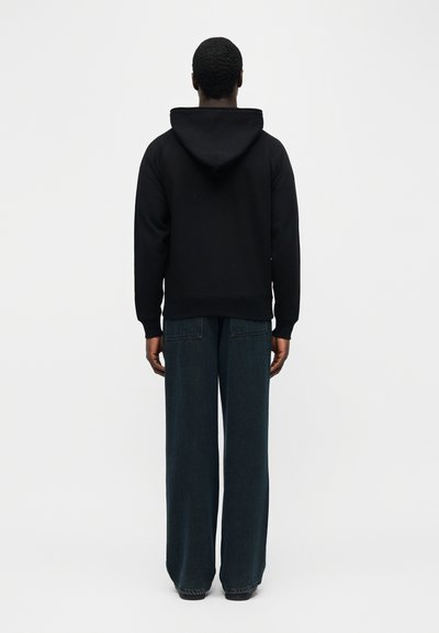 Άτομο φαίνεται από πίσω, φοράει μαύρο φούτερ με κουκούλα και φαρδιά σκούρα μπλε τζιν, μπροστά σε ένα απλό φωτεινό φόντο.