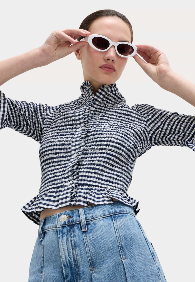 Woman adjusting white oval sunglasses, wearing a black-and-white checkered ruched blouse and high-waisted blue jeans.