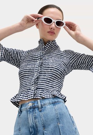 Vrouw die witte ovale zonnebril rechtzet, gekleed in een zwart-wit geruite gerimpelde blouse en een hoge taille blauwe spijkerbroek.