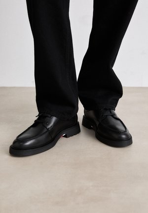 Schwarze Schnürlederschuhe, getragen zu schwarzer Hose, stehend auf hellem Boden.