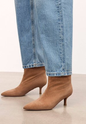 Hnědé semišové kotníkové boty se špičatou špičkou a tenkým podpatkem, v kombinaci s krátkými modrými džínovými kalhotami. Hladká textura, přiléhavý střih.