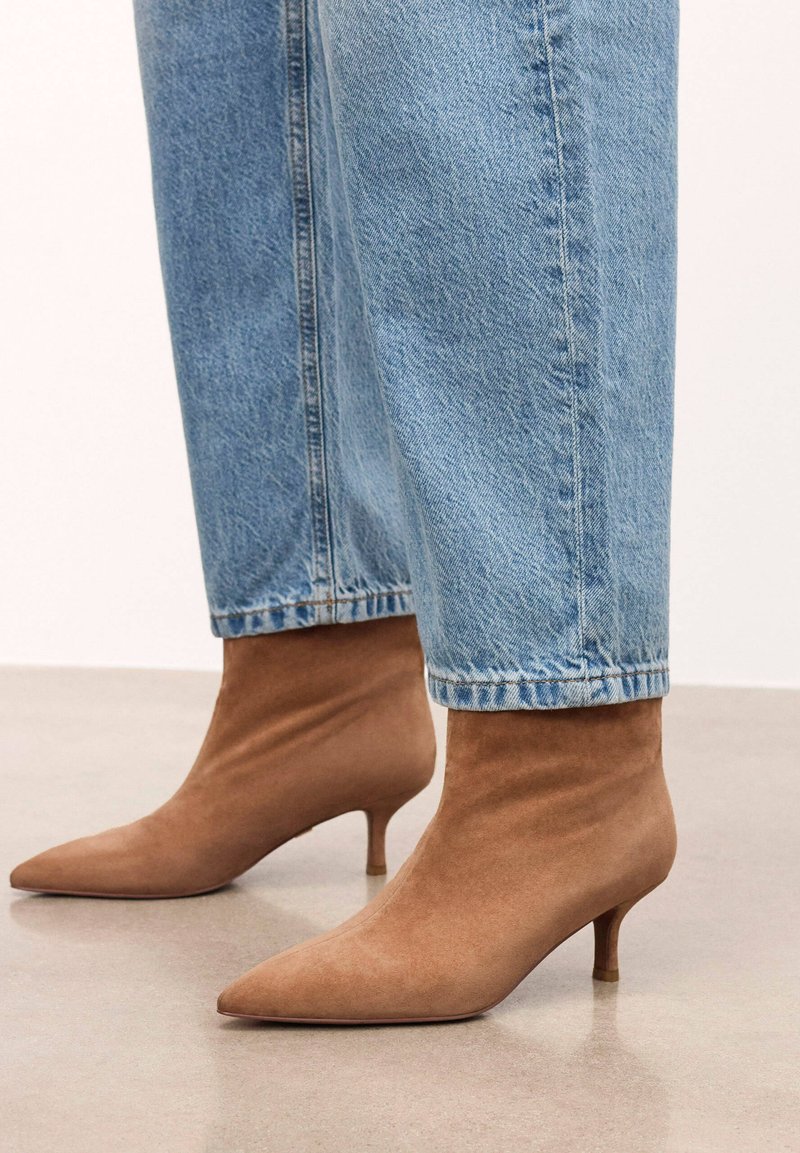 Botines de ante marrón con punta afilada y tacón delgado, combinados con jeans vaqueros azules recortados. Textura suave, diseño ajustado.